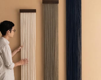 May include: Three vertical wall hangings with a minimalist design. Each piece features a dark wood top from which numerous strands of material cascade downwards. The colors are off-white, taupe, and navy blue. The wall behind the hangings is a light tan color.