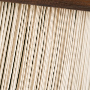 May include: Close-up of a wall hanging made of many thin, white cords hanging from a wooden bar.
