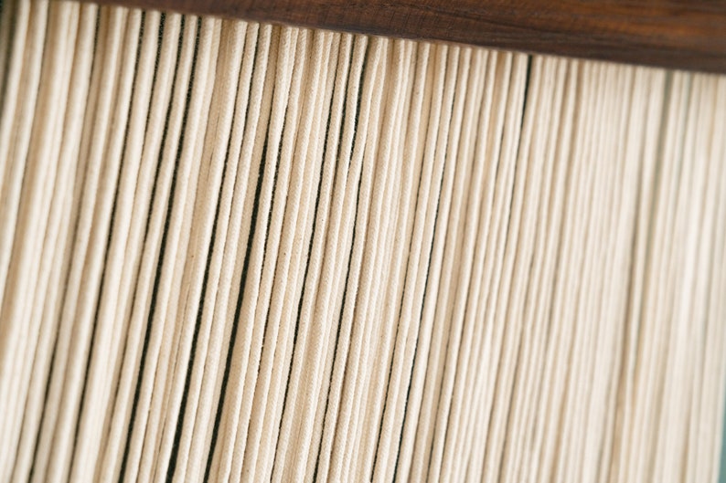 May include: Close-up of a wall hanging made of many thin, white cords hanging from a wooden bar.