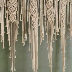 May include: A white macrame wall hanging with a woven pattern and long fringe. The hanging is made of natural cotton cord and has a bohemian style.