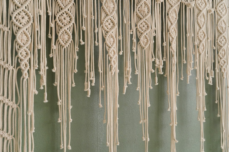 May include: A white macrame wall hanging with a woven pattern and long fringe. The hanging is made of natural cotton cord and has a bohemian style.