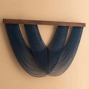 May include: A modern wall hanging featuring a dark wood beam with multiple strands of navy blue string cascading downwards. The strings are arranged in a symmetrical, flowing pattern, creating a visually appealing abstract design. This piece adds a touch of contemporary art to any space.