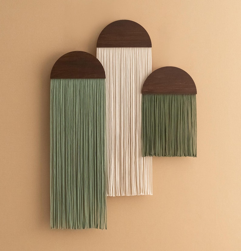 May include: Three wall hangings with wooden half-circle tops and long strands of yarn. The largest hanging has cream-colored yarn, the middle hanging has green yarn, and the smallest hanging has green yarn.