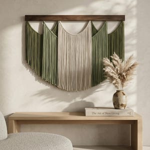 May include: A macrame wall hanging with a wooden bar, featuring olive green and beige fringe. The wall decor is displayed above a light wood bench with a vase of dried flowers and a book titled "The Art of Slow Living."