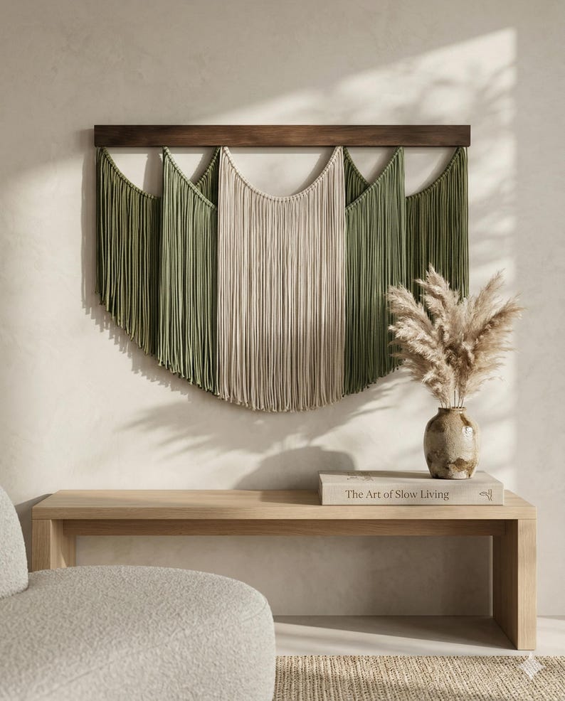 May include: A macrame wall hanging with a wooden bar, featuring olive green and beige fringe. The wall decor is displayed above a light wood bench with a vase of dried flowers and a book titled "The Art of Slow Living."