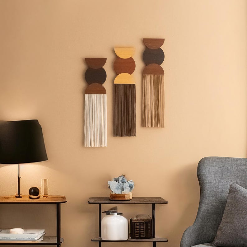 May include: Three wall hangings with a mid-century modern design. Each piece features a half-circle of wood, a circle of color, and a fringe of yarn. The color palette includes brown, beige, and cream, creating a warm, inviting aesthetic.