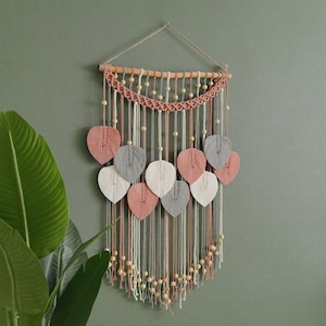 May include: A macrame wall hanging with a wooden dowel and pink, white, and gray yarn leaves. The hanging features wooden beads and a fringe.