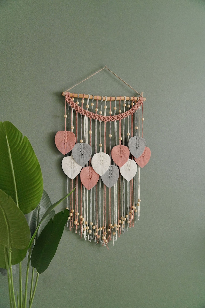 May include: A macrame wall hanging with a wooden dowel and pink, white, and gray yarn leaves. The hanging features wooden beads and a fringe.