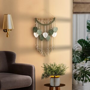 May include: A macrame wall hanging with green and white leaves, featuring a beaded fringe and a wooden dowel at the top. The wall hanging is displayed on a beige wall with a gold lamp and a potted plant.