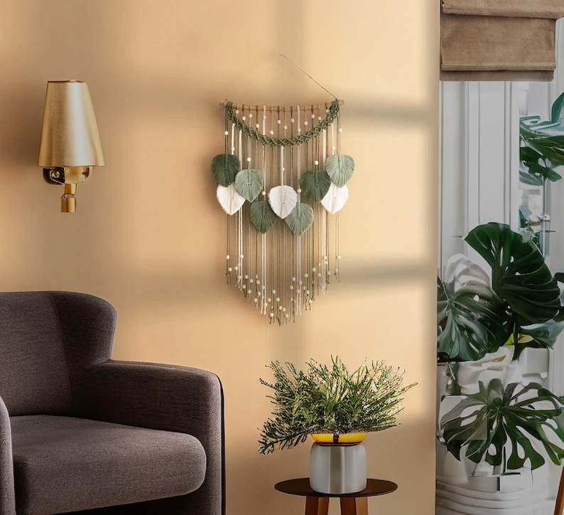 May include: A macrame wall hanging with green and white leaves, featuring a beaded fringe and a wooden dowel at the top. The wall hanging is displayed on a beige wall with a gold lamp and a potted plant.
