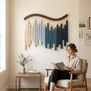 May include: A wall hanging featuring a curved dark wood bar with hanging cotton cords in an ombre color gradient. The cords transition from cream to light blue, teal, and dark navy blue, accented with small wooden beads at the top of each tassel.