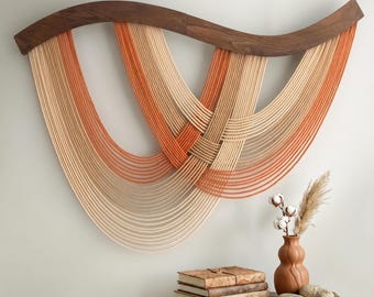 May include: A wall hanging featuring a curved dark wood top bar with multiple draped strands of rope in shades of cream, beige, and burnt orange. The rope strands are layered and intertwined to create a series of deep, overlapping U-shaped curves.