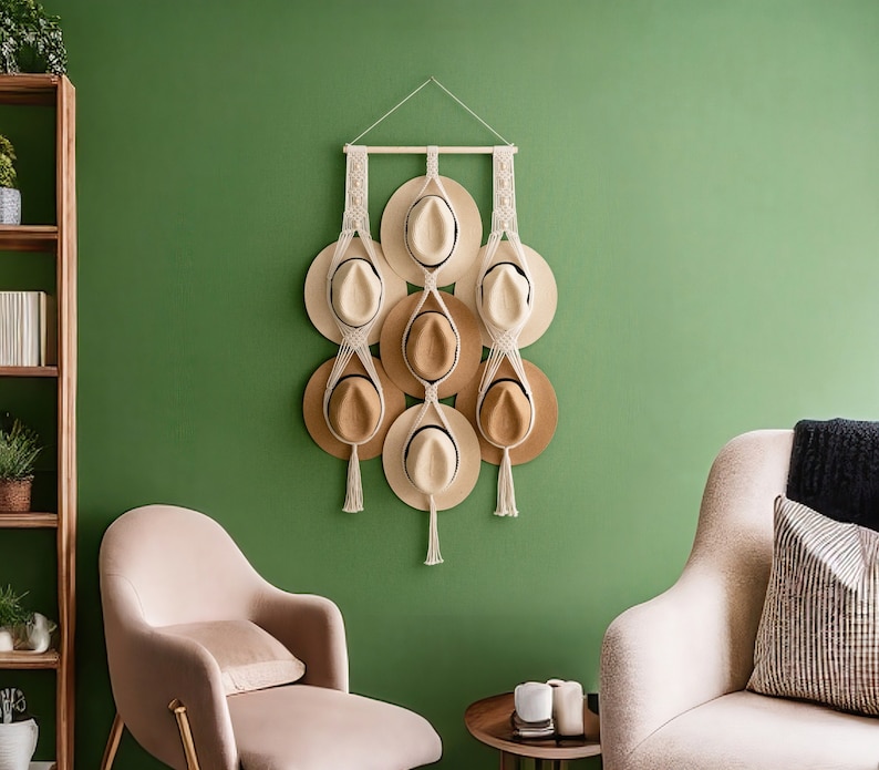 May include: A macrame hat rack displays eight hats in varying shades of tan and beige against a green wall. The rack is made of natural-colored rope and wood, with decorative tassels. The hats are arranged in a vertical pattern, creating a stylish storage solution.