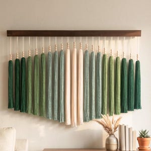 May include: A wall hanging featuring a dark brown wooden horizontal bar with multiple hanging cords. Each cord is adorned with small wooden beads and ends in a long tassel. The tassels transition in color from dark green to light green to cream.