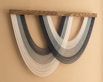 May include: A wall hanging featuring a wooden beam with two cascading, layered fiber art pieces. The fiber art transitions from white to gray to navy blue. The artwork is mounted on a beige wall.