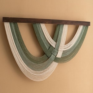 Puede incluir: Un tapiz de pared hecho de macramé tejido con un esquema de color verde y blanco. El macramé está colgado de una barra de madera oscura.