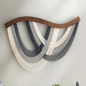 May include: A modern wall hanging featuring a curved, dark wood top with cascading strands of yarn in shades of white, gray, and navy blue. The yarn is woven together in the center, creating a unique, textured design. This piece is a stylish addition to any home decor.