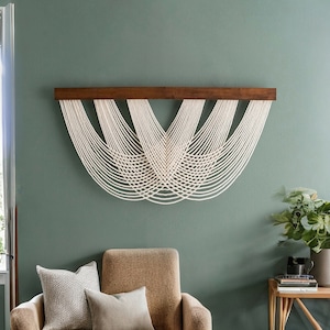 May include: A modern wall hanging featuring a dark brown wooden beam with multiple strands of off-white rope cascading downwards in a curved pattern. The wall is painted a muted green, and the room includes a chair and side table.