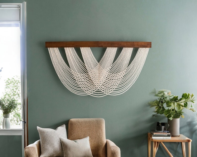May include: A modern wall hanging featuring a dark brown wooden beam with multiple strands of off-white rope cascading downwards in a curved pattern. The wall is painted a muted green, and the room includes a chair and side table.