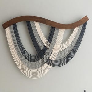 May include: A modern wall hanging featuring a curved walnut wood top and cascading strands of white, gray, and navy blue cords. The cords are woven together in the center, creating a unique, abstract design. This piece adds a touch of contemporary art to any space.