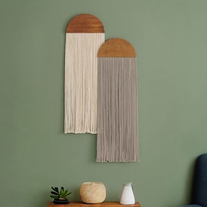 May include: Two wooden wall hangings with fringe. The left hanging has white fringe and the right hanging has gray fringe. The hangings are against a sage green wall.