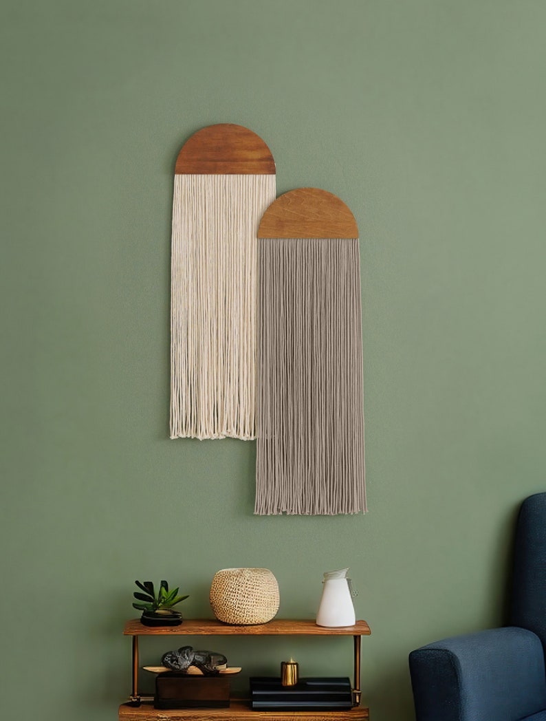 May include: Two wooden wall hangings with fringe. The left hanging has white fringe and the right hanging has gray fringe. The hangings are against a sage green wall.