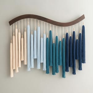 May include: A wall hanging featuring a curved dark wood bar from which hang multiple cotton yarn tassels in an ombre color gradient. The tassels transition from cream to light blue, teal, and dark navy blue, each accented with small wooden beads.
