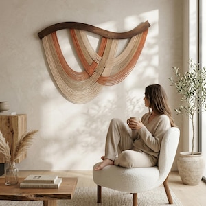 May include: A woven wall hanging with a dark brown wooden top and cascading strands in shades of beige and orange. The artwork is displayed above a white chair in a neutral-toned living room.