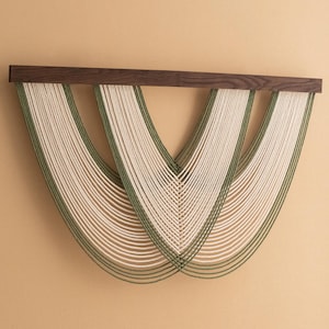 May include: A wall hanging made of woven natural fiber rope in cream and green. The rope is woven in a pattern of curved lines, creating a modern and abstract design. The hanging is mounted on a dark wood frame.