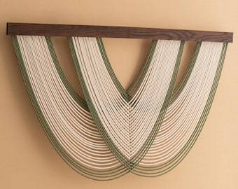 Könnte beinhalten: Ein Wandbehang aus gewebtem Naturfaserseil in Creme und Grün. Das Seil ist in einem Muster aus geschwungenen Linien gewebt, wodurch ein modernes und abstraktes Design entsteht. Der Behang ist auf einem dunklen Holzrahmen montiert.