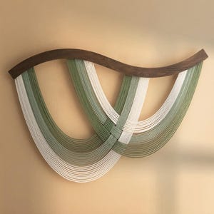 May include: A wall hanging made of woven white and green yarn, with a curved wooden frame. The yarn is woven in a pattern of overlapping loops, creating a unique and modern design.