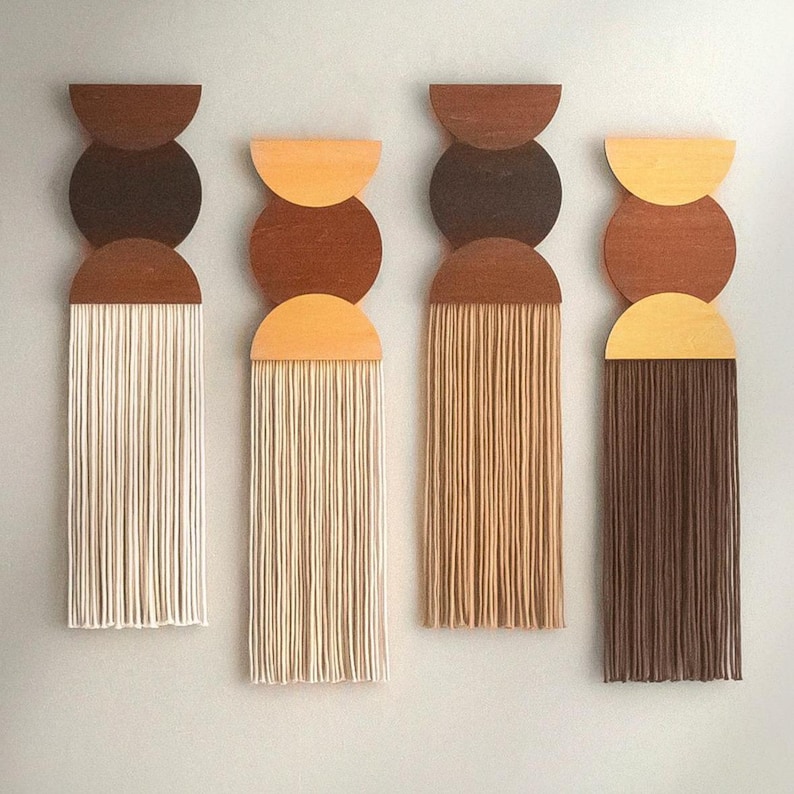 May include: Four abstract wall hangings with a mid-century modern design. Each piece features a wooden half-circle and circle in shades of brown and tan, with vertical fringe in cream, tan, and brown. A minimalist, geometric wall art.
