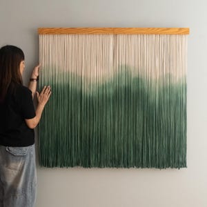 May include: A large, rectangular wall hanging with a wooden top. The hanging features vertical strands of yarn, transitioning from off-white to a deep green. The gradient effect creates a visual depth, resembling a landscape.