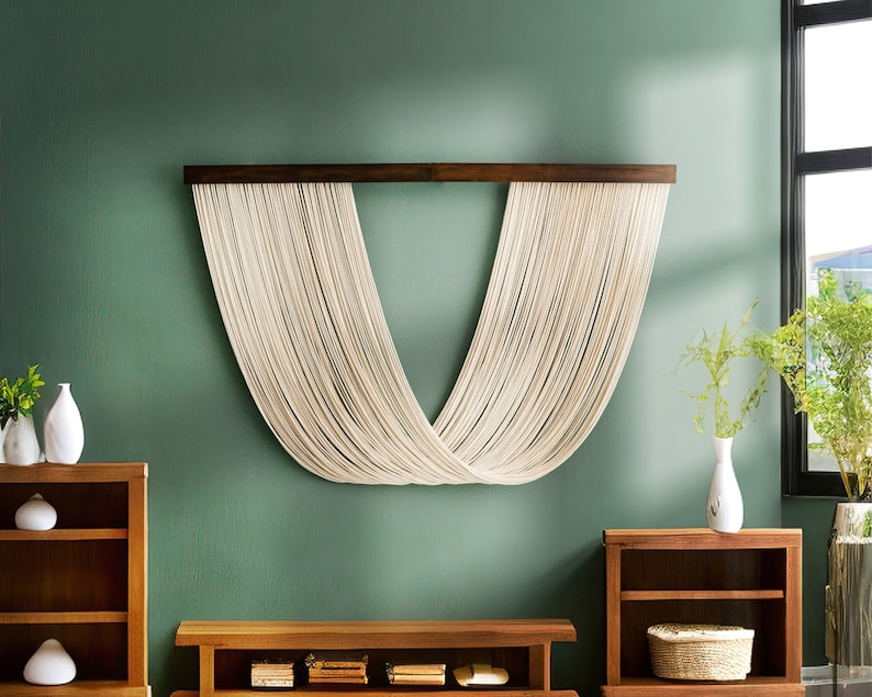 May include: A white macrame wall hanging with a wooden dowel rod, hanging on a green wall. The macrame is in the shape of a V, with the strings crossing in the middle.