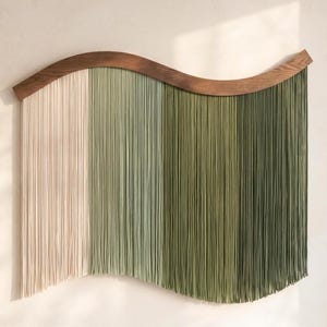 Könnte beinhalten: Eine dekorative Wandbehänge mit einem geschwungenen Oberteil aus Holz und vertikalen Strängen in einem Farbverlauf. Die Farben gehen von Off-White in Grüntöne über und erzeugen ein strukturiertes, fließendes Design. Das Oberteil aus Holz fügt ein natürliches Element hinzu.