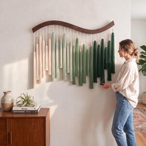 May include: A wall hanging featuring a curved dark wood bar with hanging cotton cords in an ombre color gradient. The cords transition from cream to sage green and dark forest green, accented with small wooden beads above each tassel fringe.