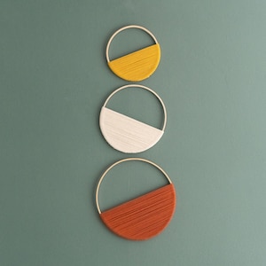 Puede incluir: Tres colgantes de pared circulares con marco de madera y relleno de hilo. El superior es amarillo, el central blanco y el inferior naranja. El hilo llena la mitad de cada círculo, creando un diseño de bloque de color diagonal.