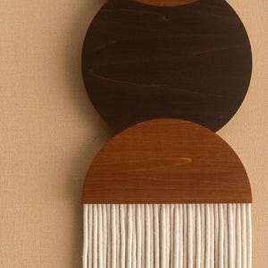 May include: Wooden wall art featuring three circular and semi-circular elements in shades of brown. The top two are solid wood, while the bottom element has a fringe of white rope. The background is a neutral beige.
