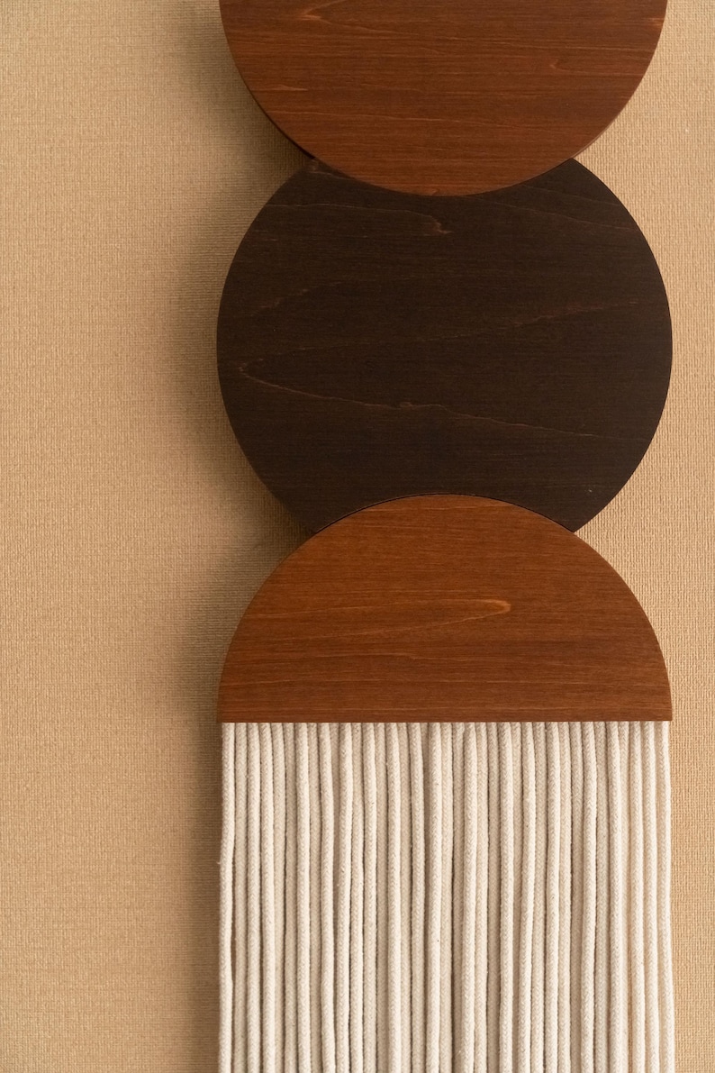 May include: Wooden wall art featuring three circular and semi-circular elements in shades of brown. The top two are solid wood, while the bottom element has a fringe of white rope. The background is a neutral beige.