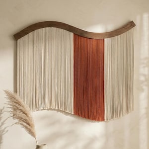 May include: A large wall hanging with a wooden curved top and vertical strands. The strands are divided into three sections: off-white, rust-orange, and off-white. The piece is a decorative element for a home.