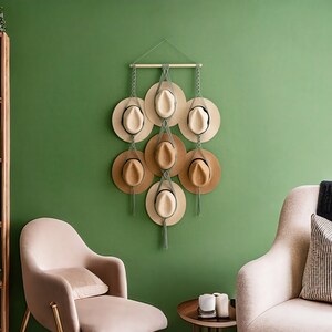 May include: A wall-mounted hat rack displaying seven hats of varying shades of tan and beige. The hats are arranged in a cascading pattern and suspended by chains and macrame. The background is a solid green wall, with two chairs and a small table in the foreground.