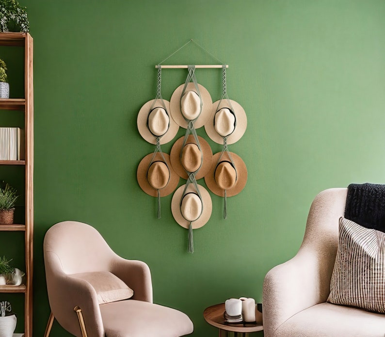 May include: A wall-mounted hat rack displaying seven hats of varying shades of tan and beige. The hats are arranged in a cascading pattern and suspended by chains and macrame. The background is a solid green wall, with two chairs and a small table in the foreground.