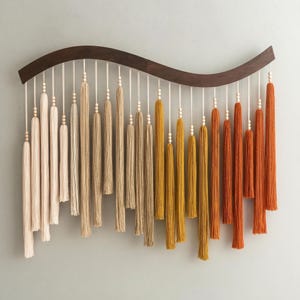 May include: A decorative wall hanging featuring a dark wood curved top with multiple tassels in varying shades of cream, tan, yellow, and orange. Each tassel is suspended by a white string and adorned with small wooden beads. The wall art is designed to add a touch of bohemian style.