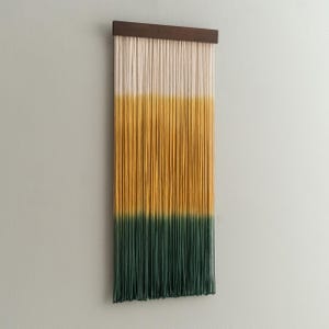 May include: A wall hanging with a wooden top and vertical strands in cream, yellow, and green. The strands are arranged in a gradient, transitioning from cream to yellow to green, creating a decorative piece for home decor.