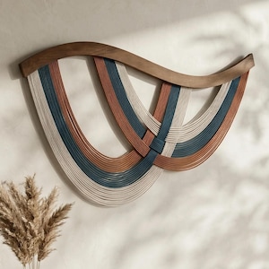 Könnte beinhalten: Ein Wandbehang mit einem geschwungenen Rahmen aus dunklem Holz und mehreren Seilsträngen in den Farben Creme, Terrakotta und Marineblau. Die Seile sind in überlappenden, U-förmigen Mustern angeordnet, die ein geschichtetes, gewebtes Aussehen erzeugen.