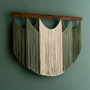 May include: A macrame wall hanging featuring a dark wooden rod and cascading strands of olive green and cream-colored cotton. The design has a symmetrical, arched shape, creating a modern, textured wall decor piece.