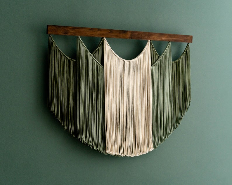 May include: A macrame wall hanging featuring a dark wooden rod and cascading strands of olive green and cream-colored cotton. The design has a symmetrical, arched shape, creating a modern, textured wall decor piece.