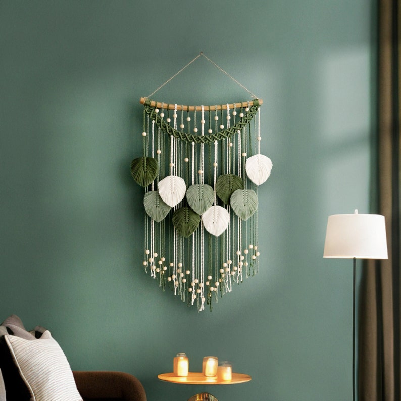 May include: A green and white macrame wall hanging with a wooden dowel and white beads. The hanging features a series of green and white leaves.