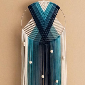 Puede incluir: Un tapiz de macramé azul y blanco con un anillo circular de madera y cuentas blancas. El macramé está hecho con una variedad de cordones azules y blancos, creando un efecto degradado.