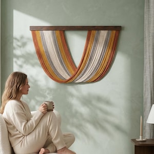 May include: A wall hanging with a dark brown wooden top and colorful yarn strands in shades of orange, yellow, gray, and cream. The yarn is arranged in a curved, draped design. The wall is painted a light green color.